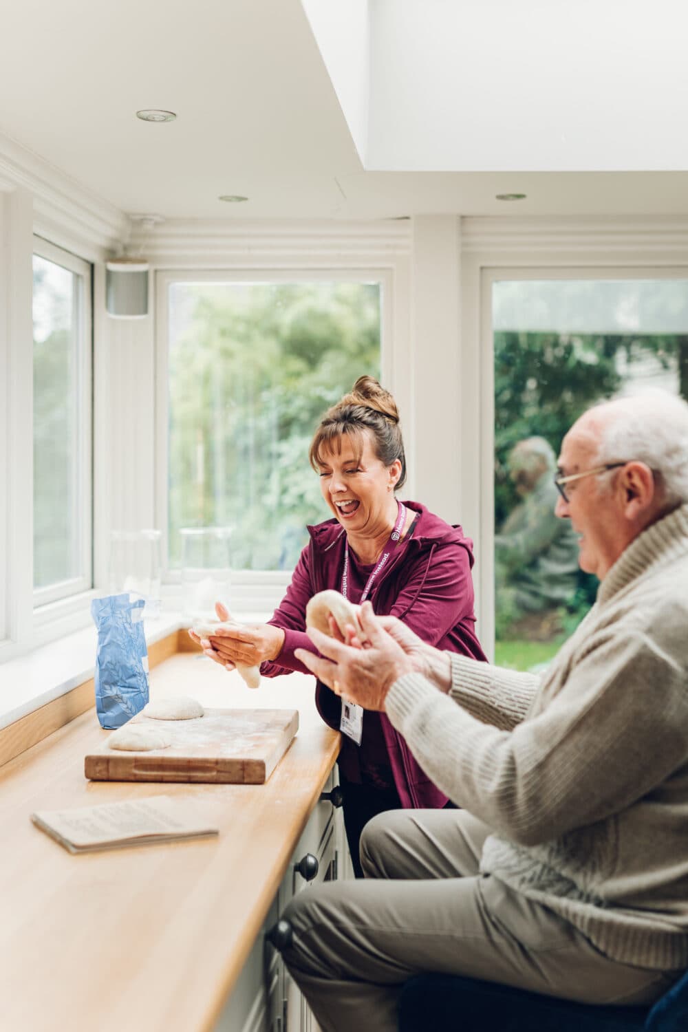 Woman and elderly man smiling and making dough together in a bright kitchen. - Home Instead