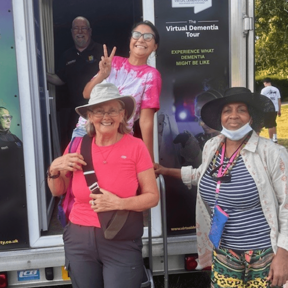 Four people smile outside a trailer with "The Virtual Dementia Tour" sign. - Home Instead