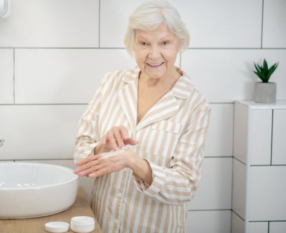 Elderly woman applying cream to her hands, smiling in a bathroom with striped pajamas. - Home Instead