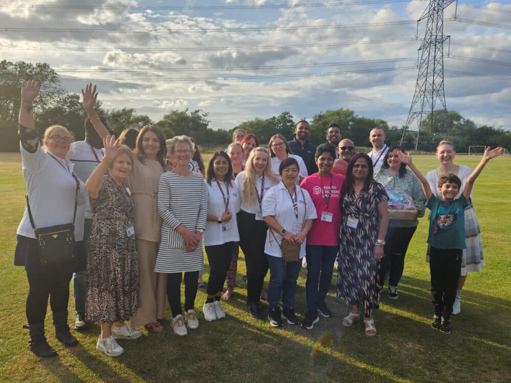 A group of people posing and smiling outdoors on a grassy field under a partly cloudy sky. - Home Instead