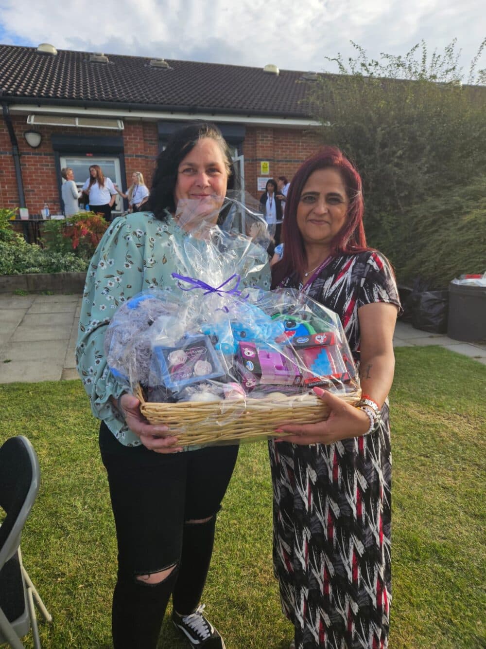 Two women smiling and holding a gift basket outdoors on a sunny day. - Home Instead