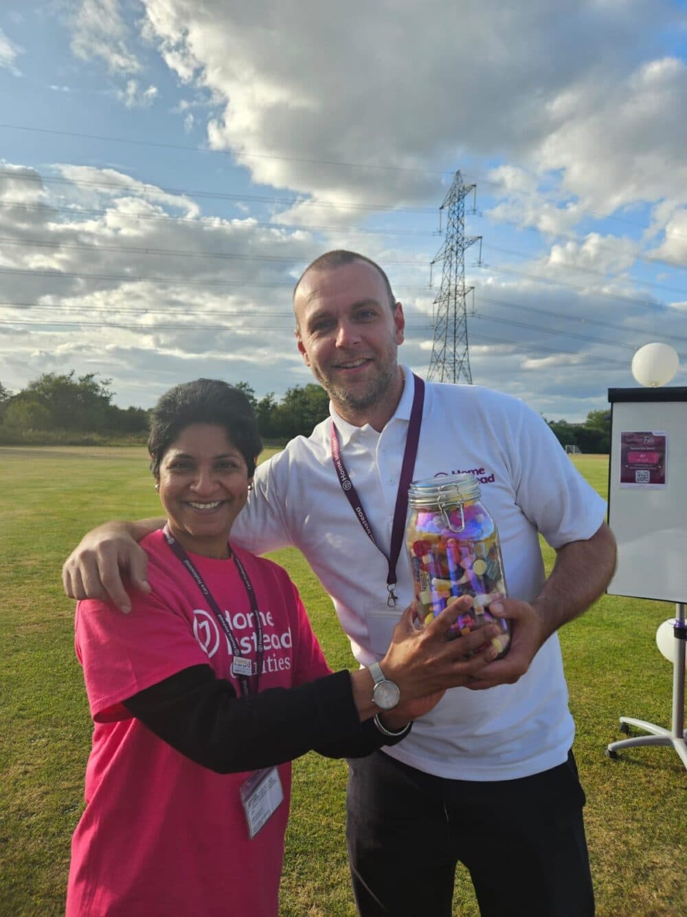 Two people standing on grass, smiling and holding a jar of sweets, with a cloudy sky in the background. - Home Instead