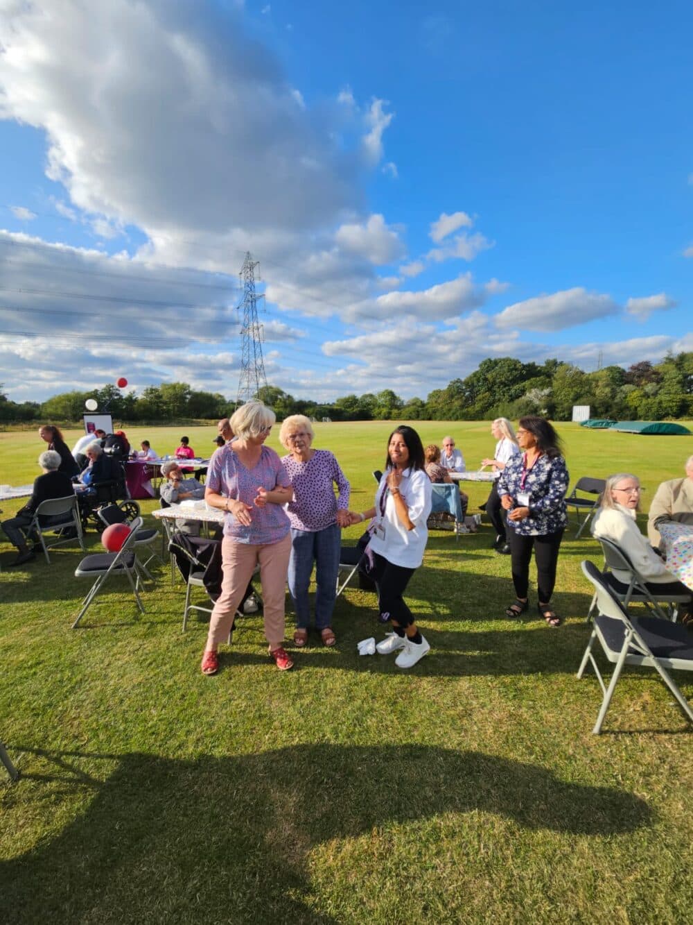 People enjoying an outdoor gathering on a sunny day, with chairs and tables set up on a grassy field. - Home Instead