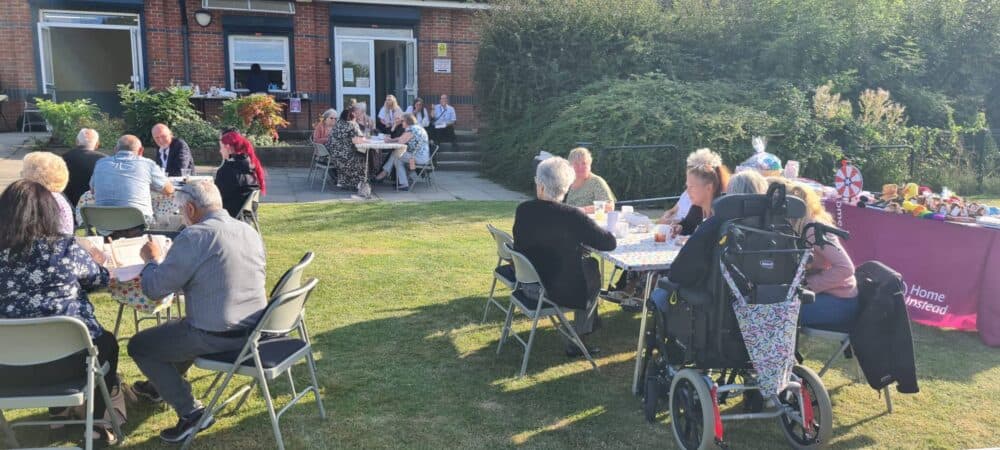 People enjoying a garden party with tables, chairs, and a booth on a sunny day. - Home Instead