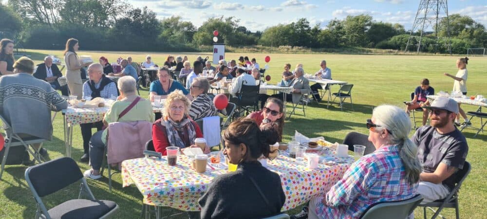 People seated at tables outdoors at a community gathering on a sunny day, with trees and a field in the background. - Home Instead