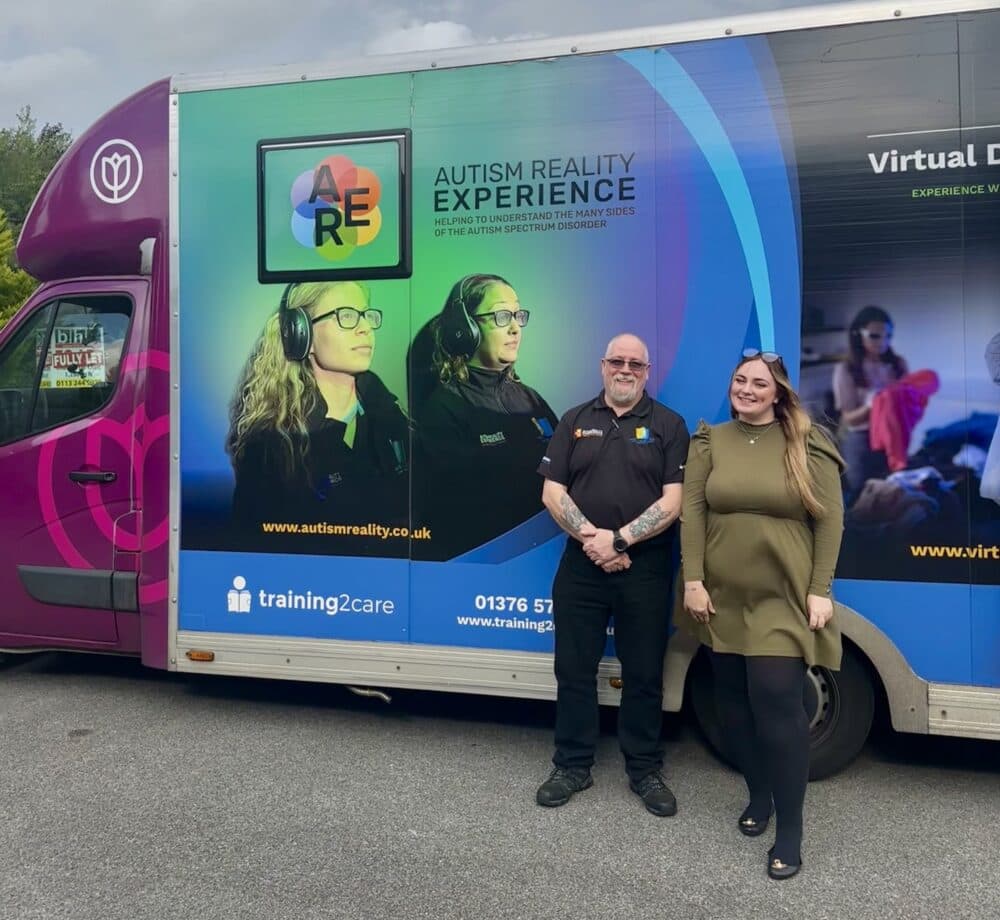 Two people stand smiling in front of the Autism Reality Experience truck with colorful graphics. - Home Instead