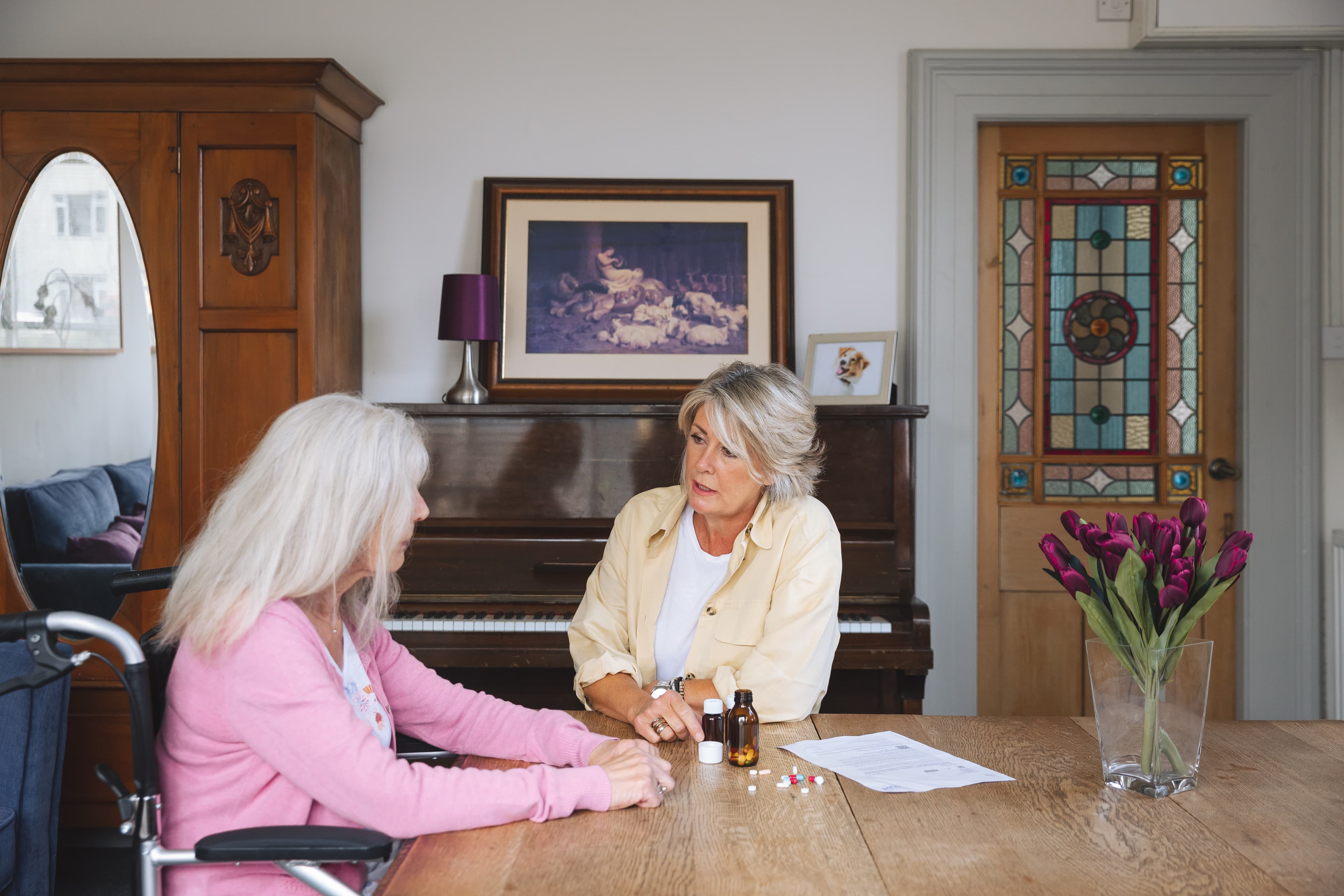 Image of daughter and mother sitting at dining table. Daughter is supporting mother with her daily medication.