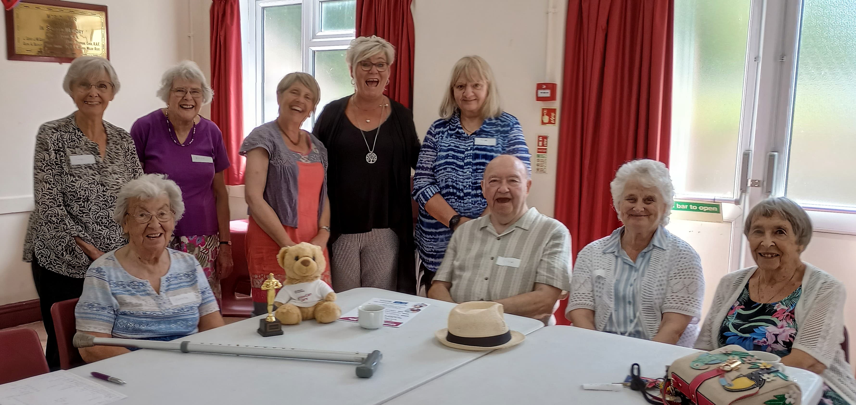 Group of smiling elderly people posing with a teddy bear in a bright room with red curtains. - Home Instead
