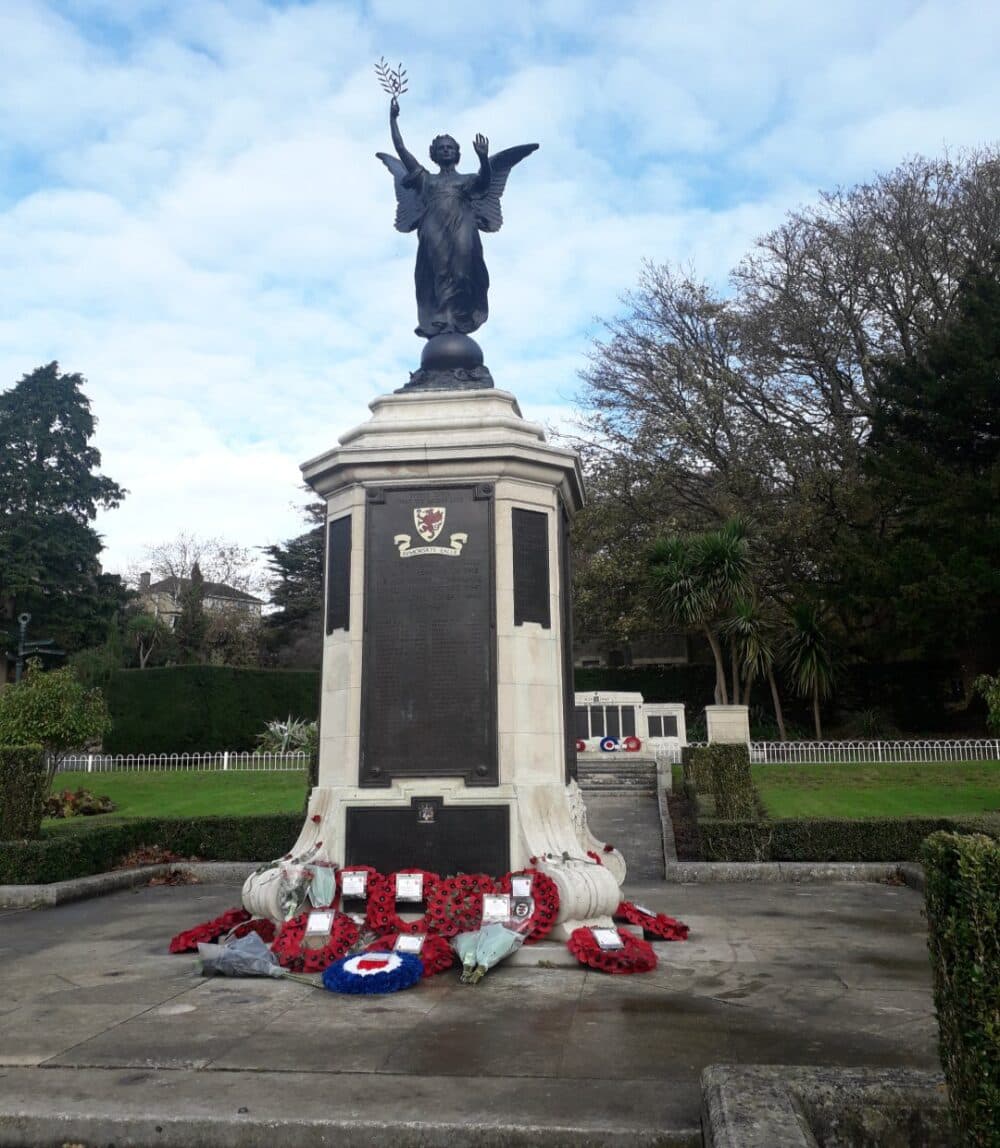 Statue atop a tall monument with wreaths at the base, set in a park with trees and a cloudy sky. - Home Instead