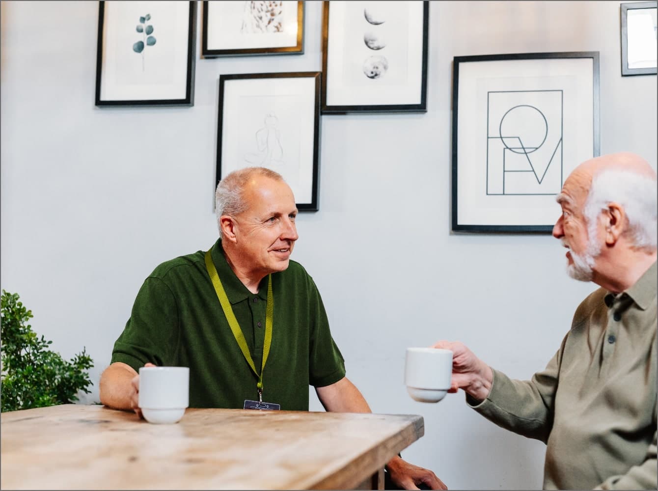 Two older men sit at a wooden table, holding mugs, with framed art on the wall behind them. Overnight Care at Home Instead North Lanarkshire and Glasgow East