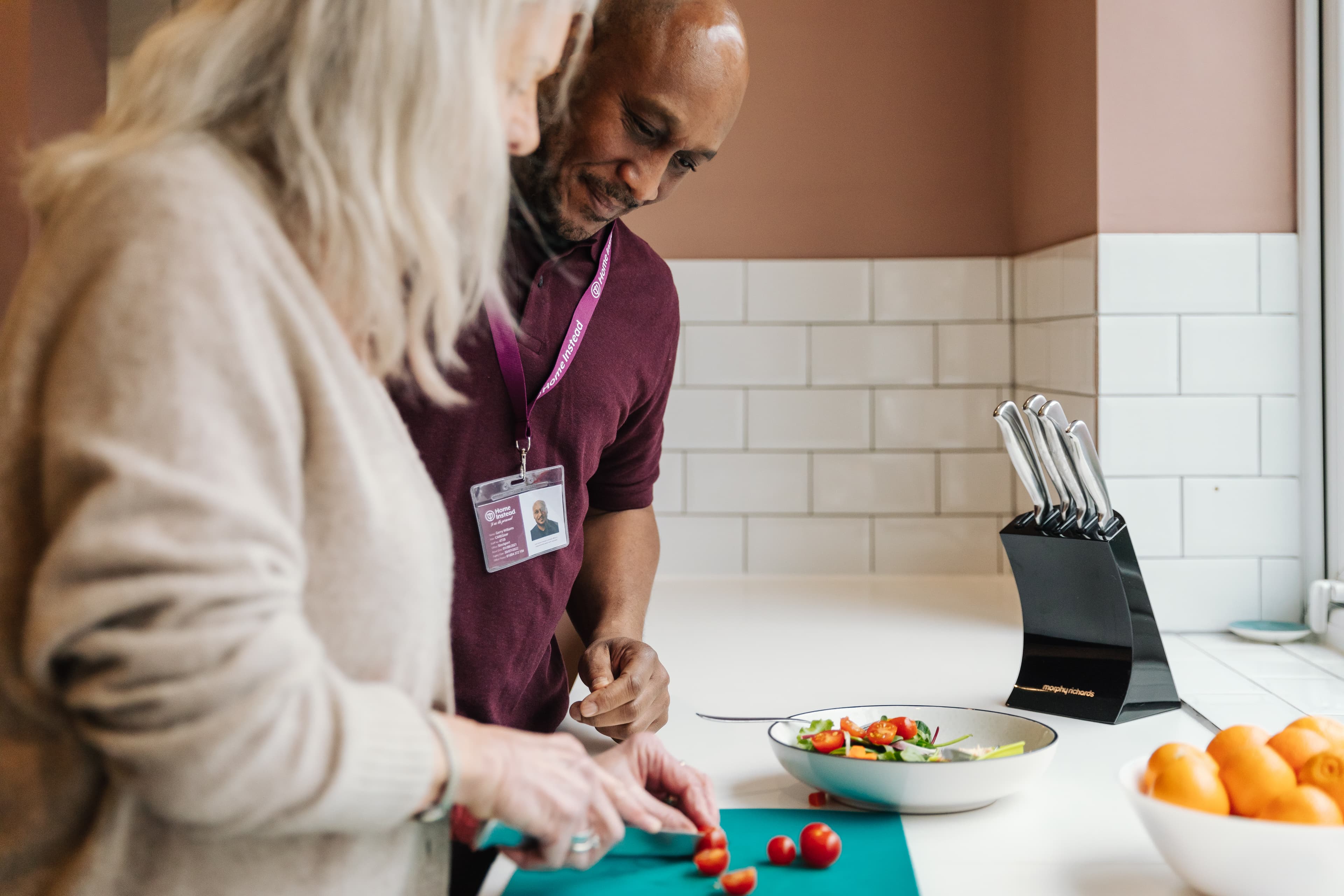 Two people slicing tomatoes in a kitchen next to a bowl of salad and a knife set. - Home Instead