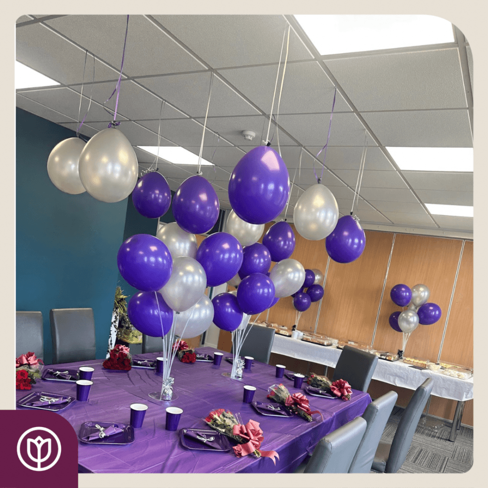 Office party setup with purple and silver balloons, purple tablecloth, and three tables arranged with decorations and chairs. - Home Instead