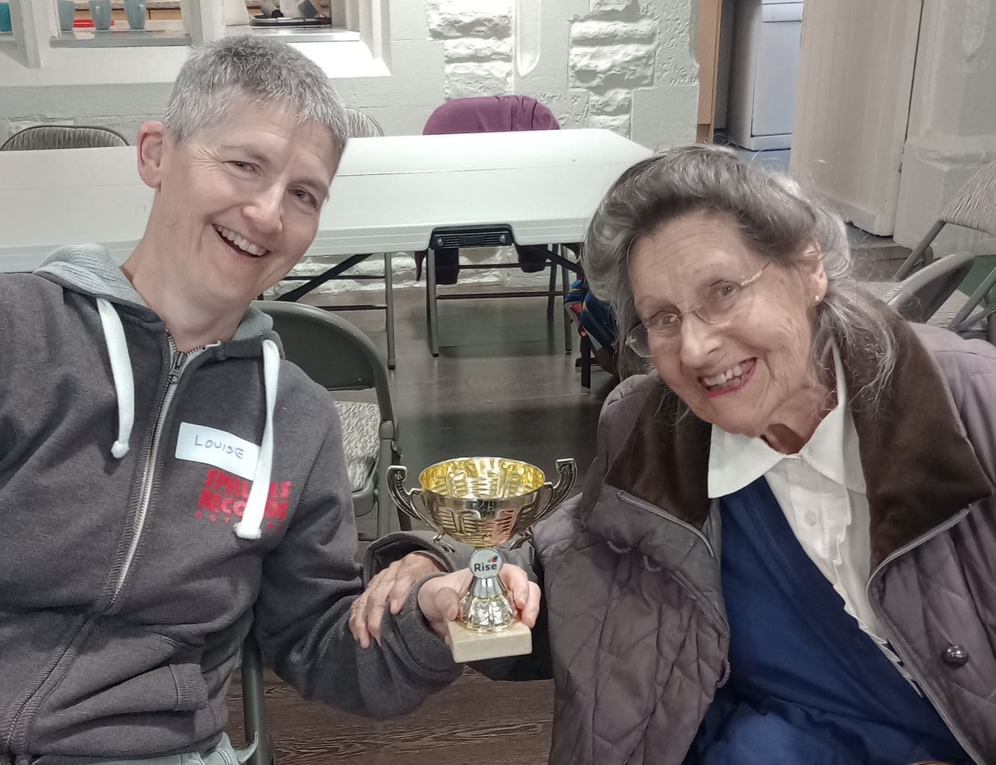 Two smiling people sitting and holding a trophy together in a room. - Home Instead