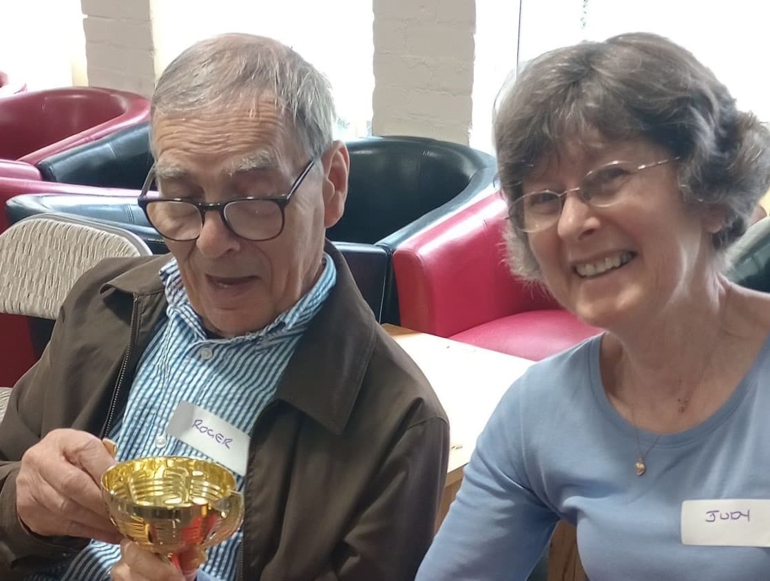 An older man and woman, both smiling, sit together; the man holds a small trophy. - Home Instead