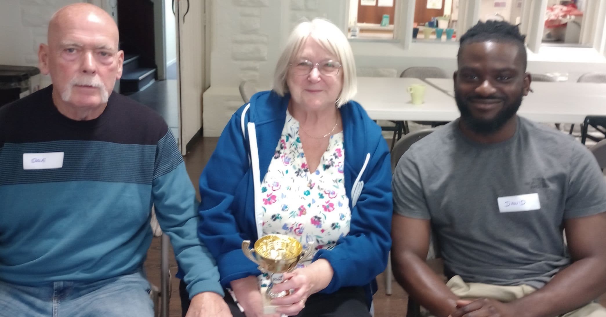 Three people sitting together, one holding a trophy, all wearing name tags and smiling at the camera. - Home Instead