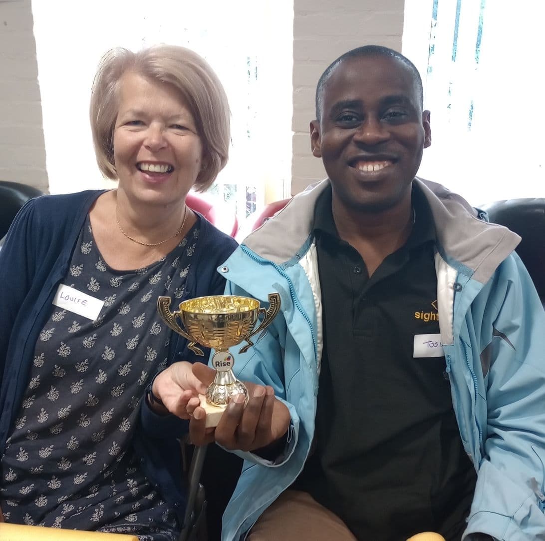 Two people smiling and holding a small trophy, sitting in a room. Both have name tags. - Home Instead