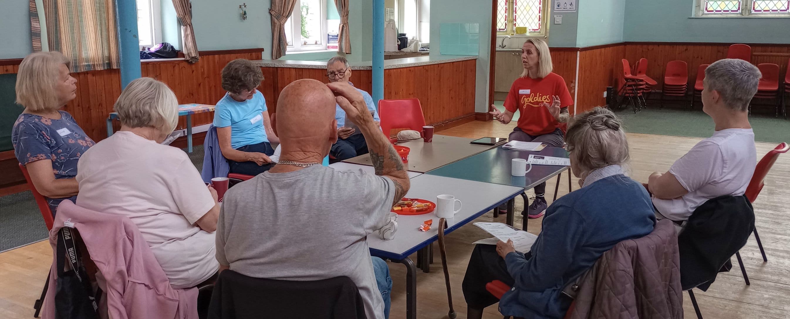 A group of people sit in a circle in a community hall, engaged in discussion with a facilitator. - Home Instead