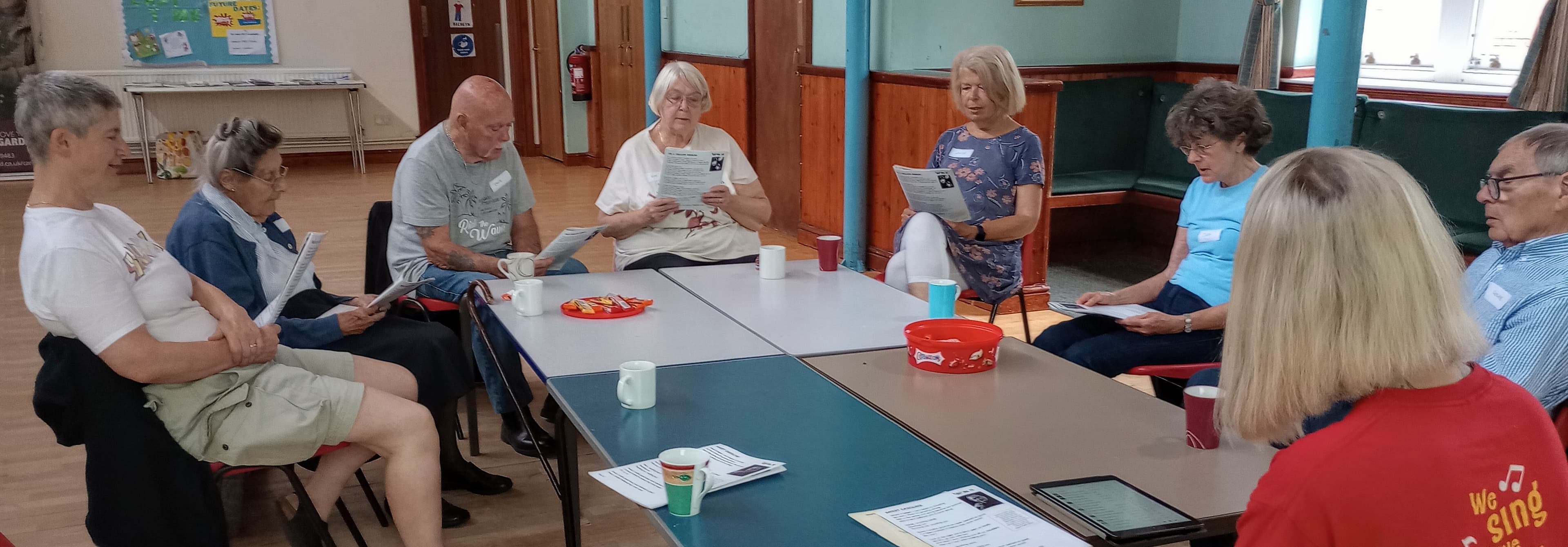A group of older adults sitting in a circle, holding papers, and engaging in a discussion in a community hall. - Home Instead