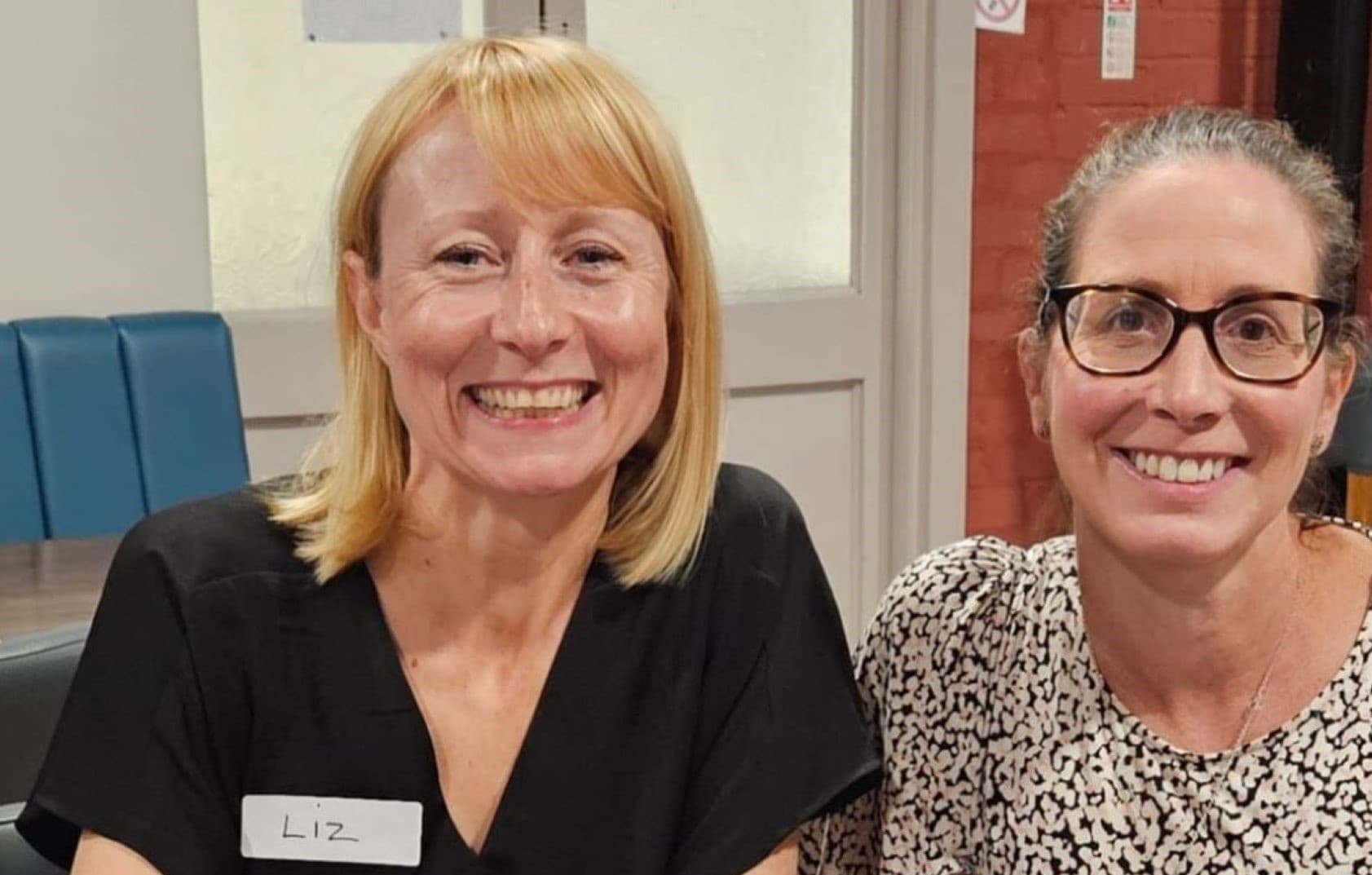 Two women smiling, one with a name tag that says "Liz," sitting indoors near a door and blue seating. - Home Instead