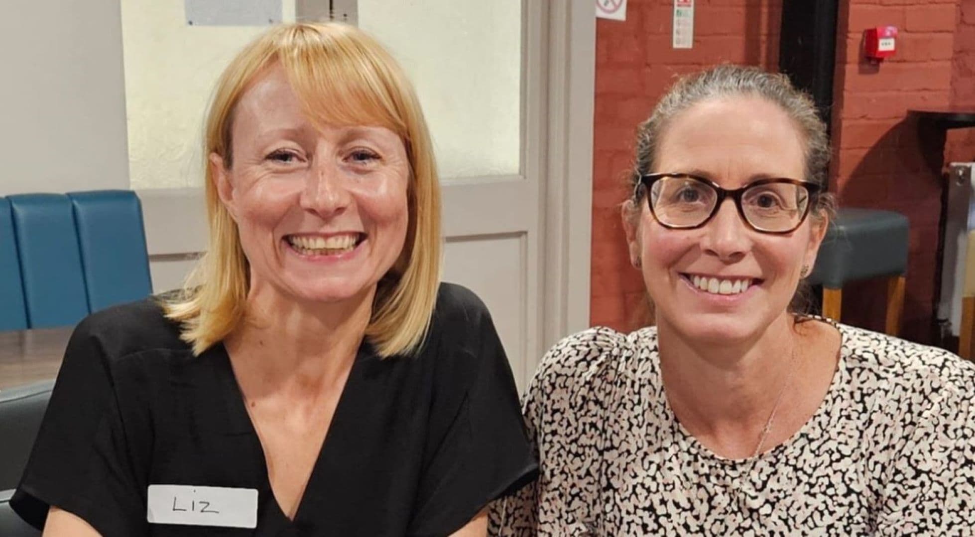 Two women smiling and sitting indoors; one wears a black shirt with a name tag, the other a patterned white top. - Home Instead