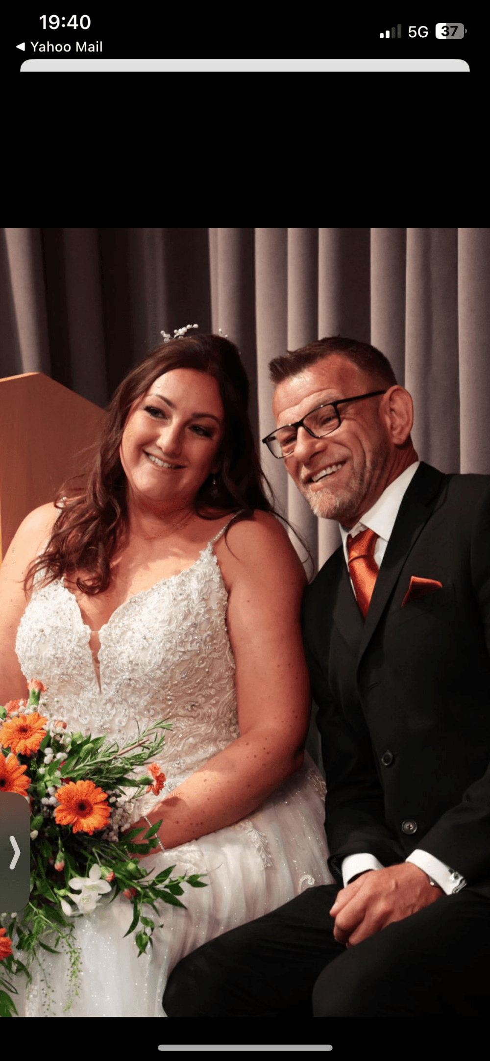A bride and groom smile while seated, holding a bouquet of orange flowers. - Home Instead