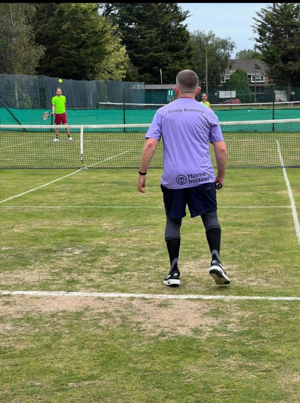 Two people playing tennis on a grass court, one preparing to serve. - Home Instead