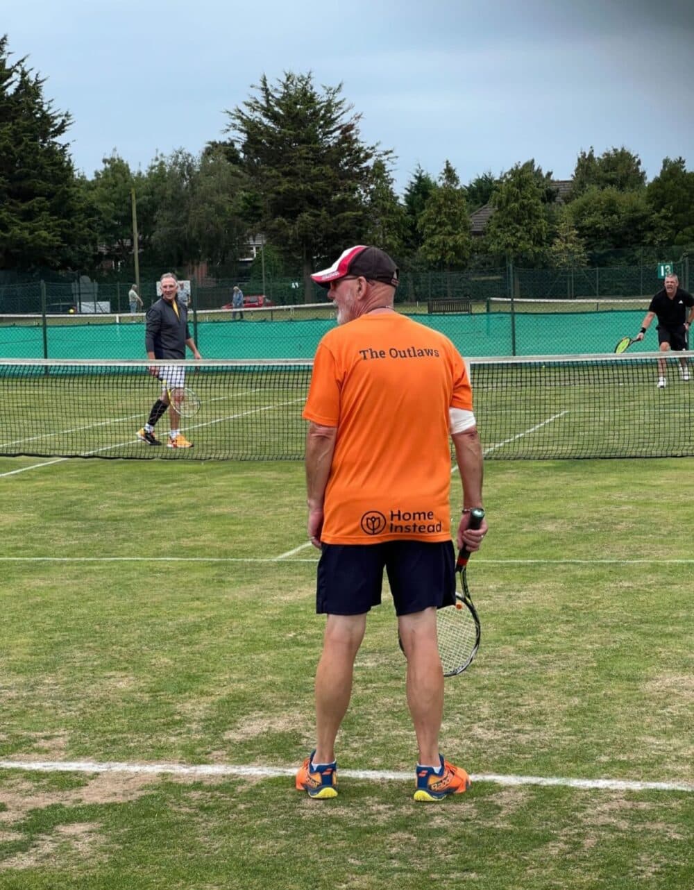 A tennis player in an orange shirt stands on a grass court, watching a doubles match. - Home Instead