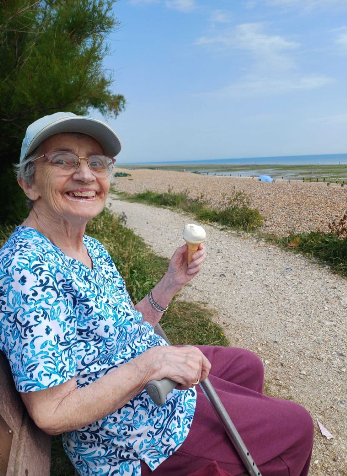 Elderly woman smiling, sitting on a bench by the beach, holding an ice cream cone, wearing a cap and patterned shirt. - Home Instead