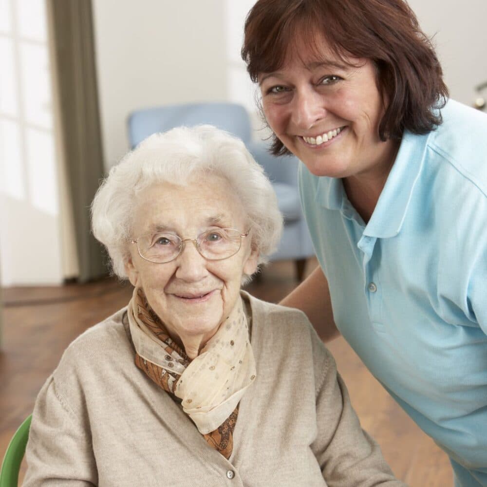 An elderly woman smiling with a caregiver in a light blue shirt, both looking at the camera. - Home Instead