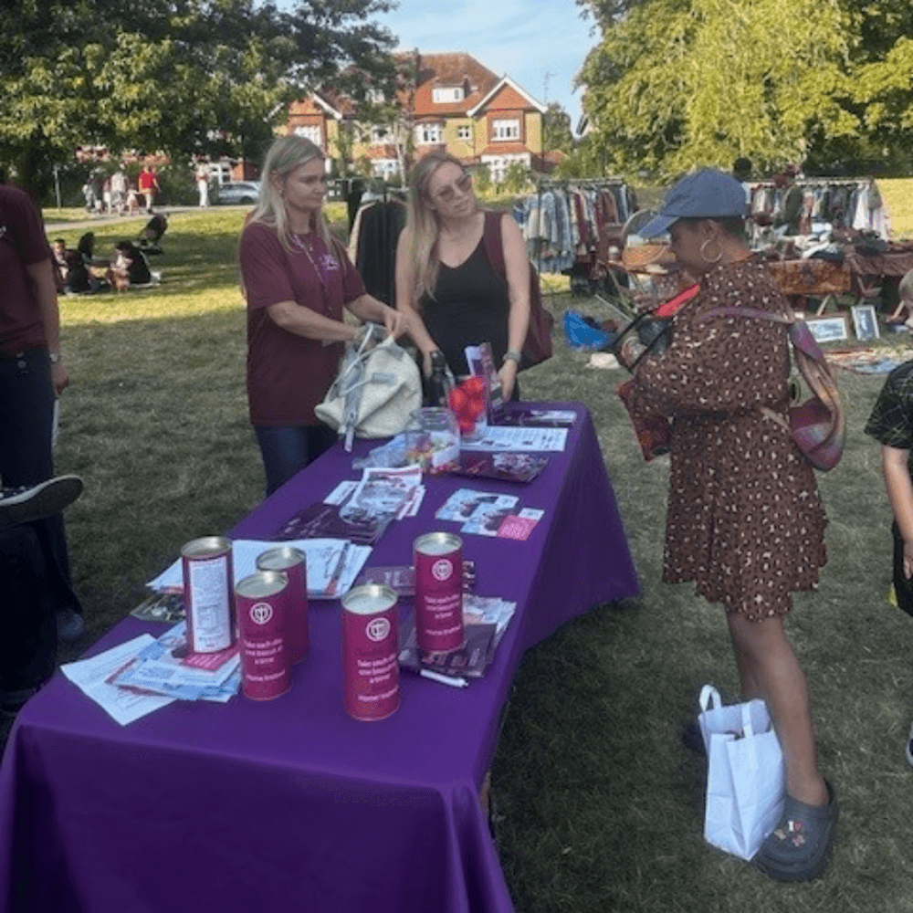 People at an outdoor fair, engaging at a table with brochures and collection tins on a purple cloth. - Home Instead