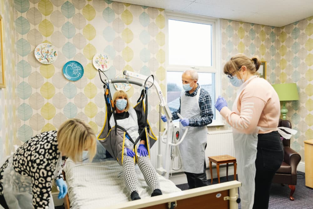 A woman being assisted onto a bed with a lift by three masked caregivers in a decorated room. - Home Instead