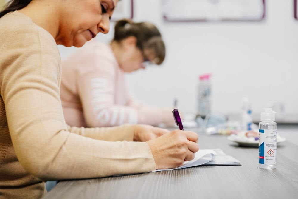 Two women sitting at a table, writing in notebooks with pens. Bottles and snacks are on the table. - Home Instead