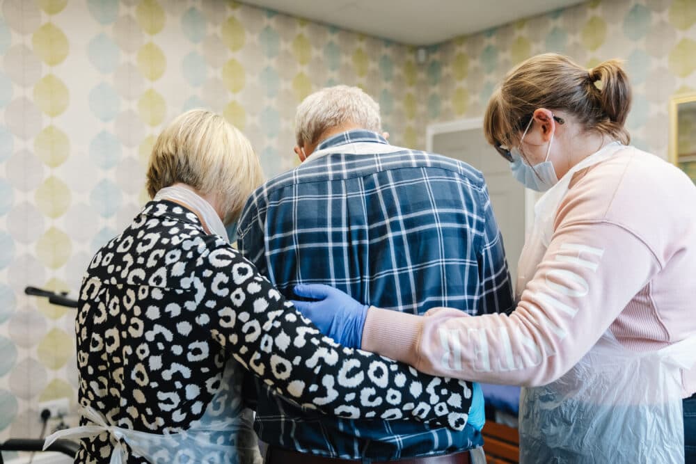Two caregivers support an elderly man as he walks in a brightly decorated room. - Home Instead