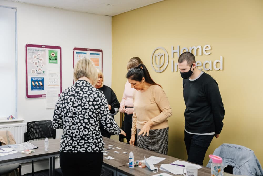 People in a meeting room gather around a table with papers and supplies at a place labeled "Home Instead. - Home Instead