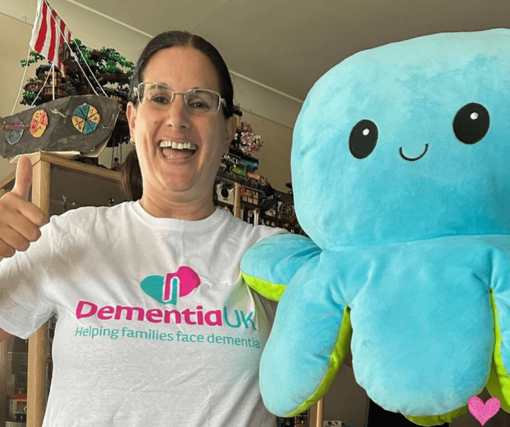 Person wearing a Dementia UK shirt, smiling and giving a thumbs-up, holds a large cute blue stuffed octopus. - Home Instead