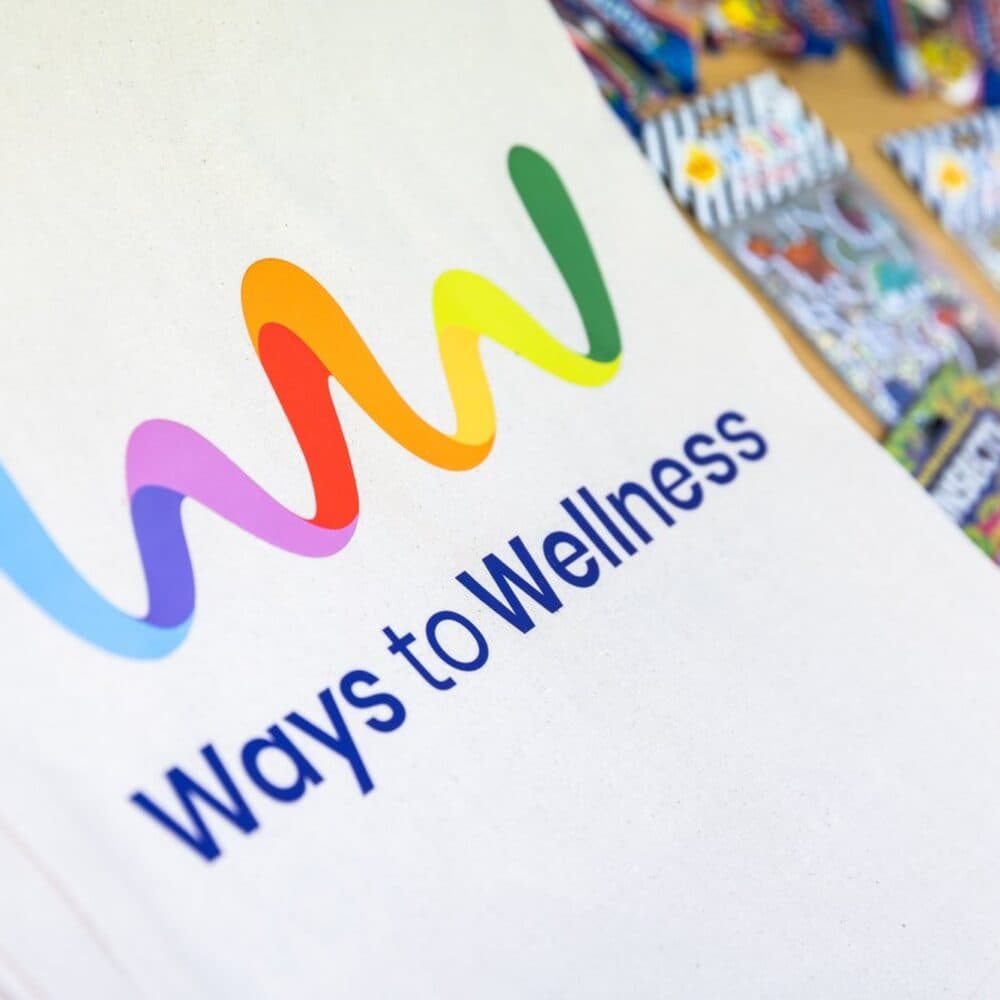 Canvas bag with a colorful wave logo and text "Ways to Wellness" surrounded by brochures. - Home Instead