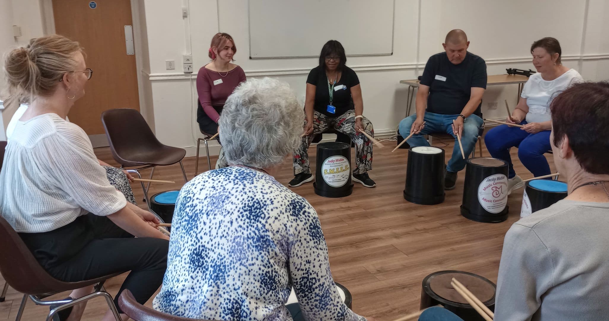 A group of people sitting in a circle, playing drums made from buckets in a classroom setting. - Home Instead