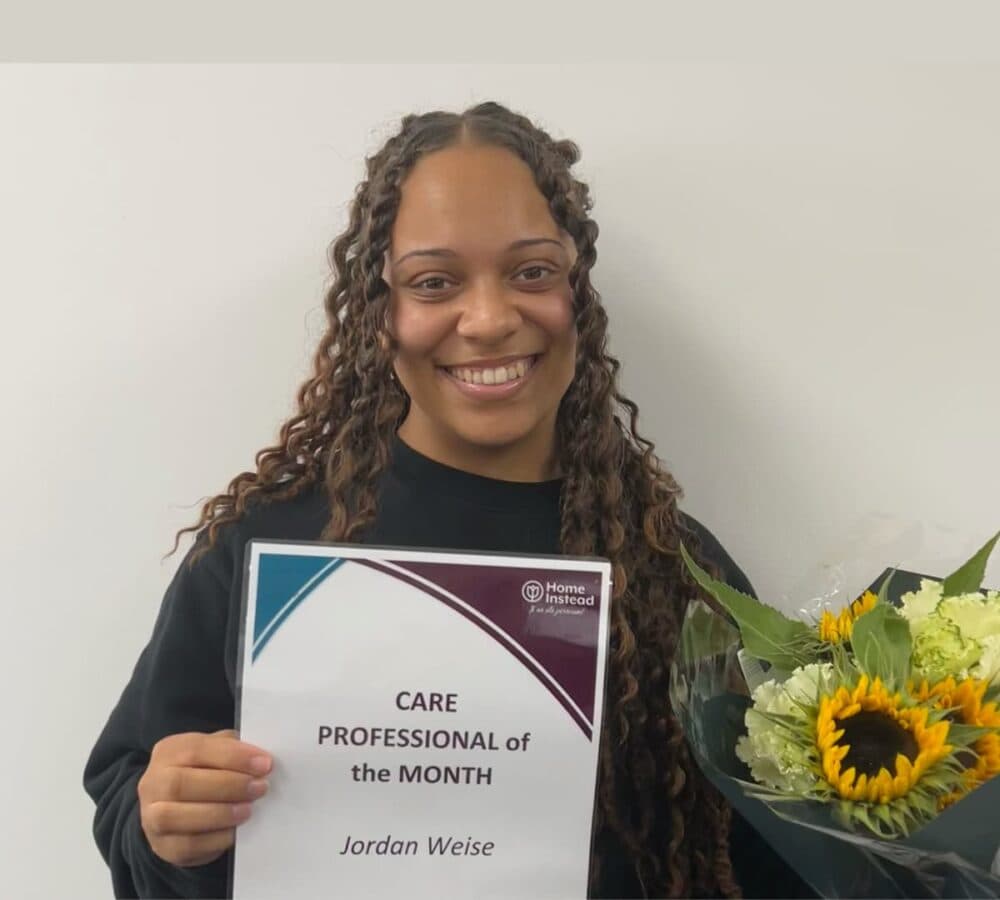 A Care Professional with curly hair smiling while holding her Care Professional of the month awards and holding a bouquet of sunflowers