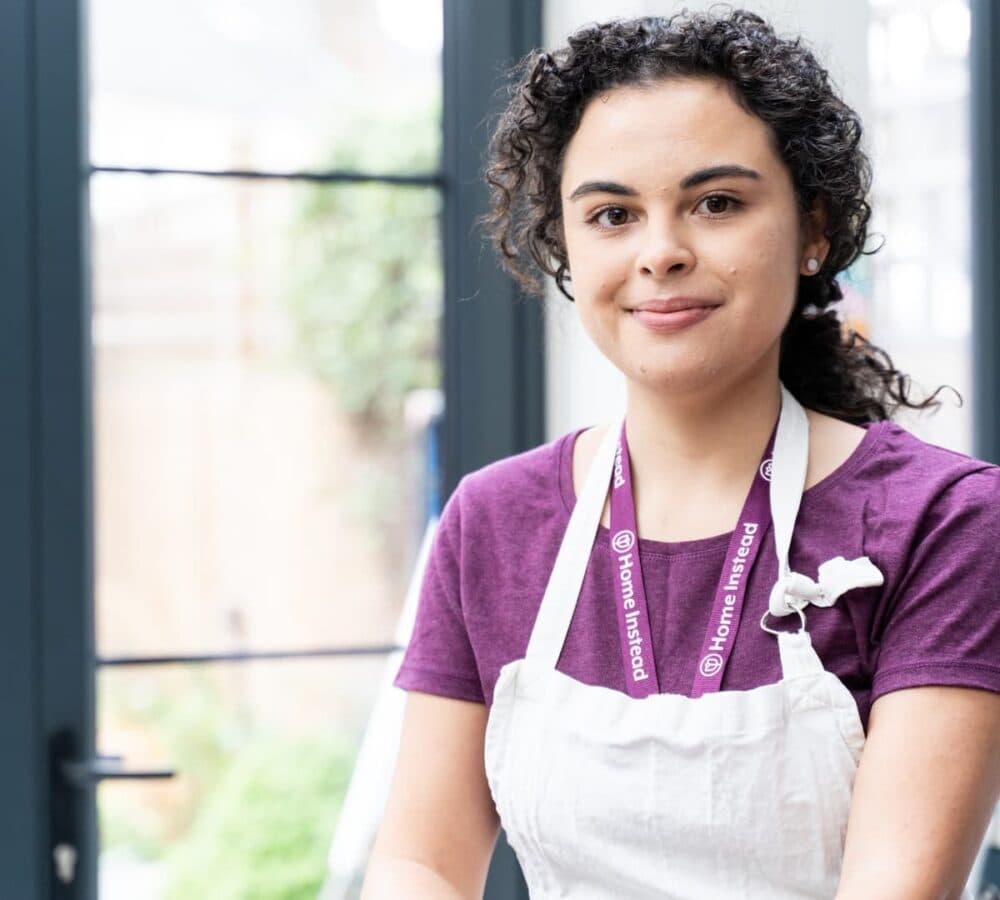 A lady with curly hair smiling and wearing an apron and maroon shirt