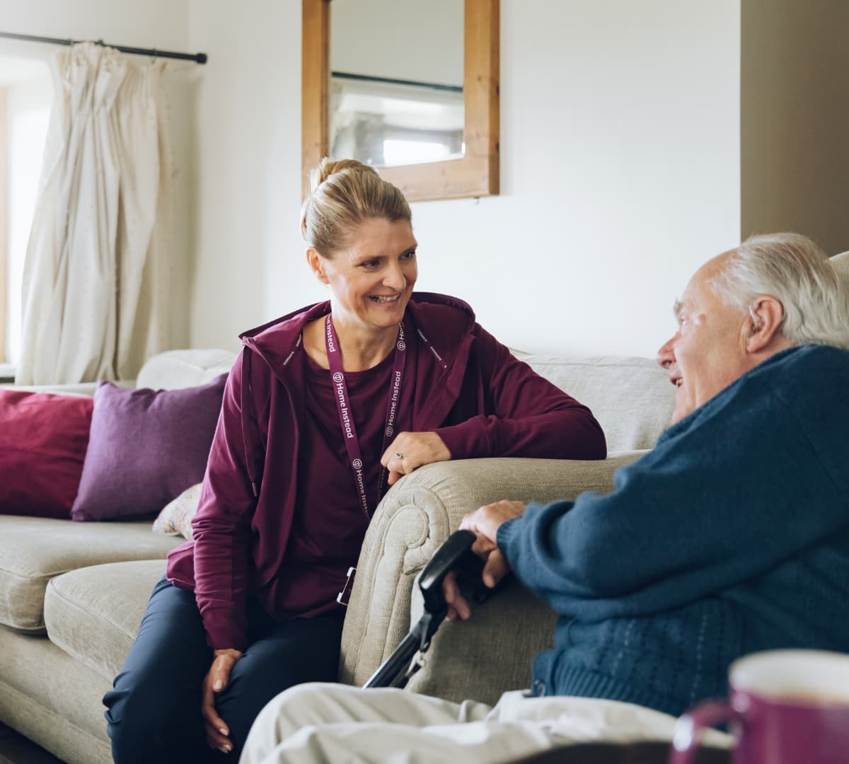 A Senior male wearing bluw whith white hair sitting on couch and holding a cane while talking to his carer wearing maroon sweater sitting on a couch and smiling inside the house