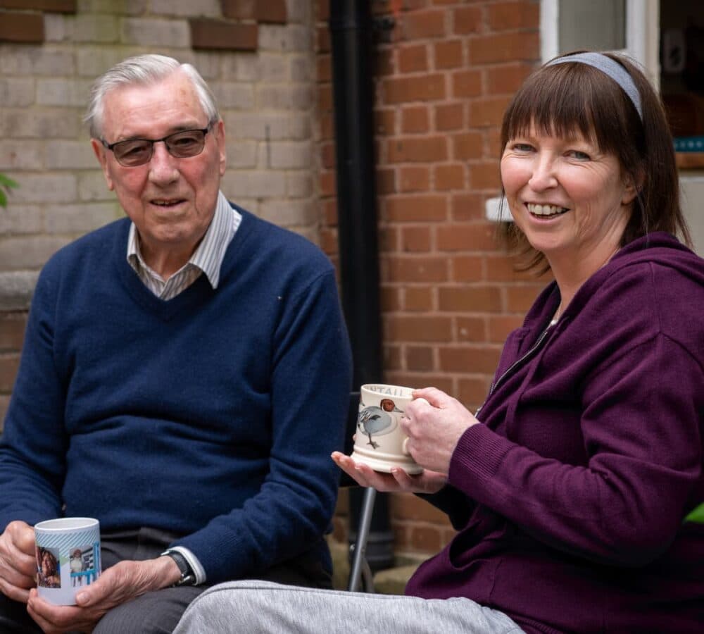 A Senior man with white hair and blue sweater wearing eyeglasses and holding a cup with his carer smiling and wearing a maroon jacket with headband while holding a cup outside the house