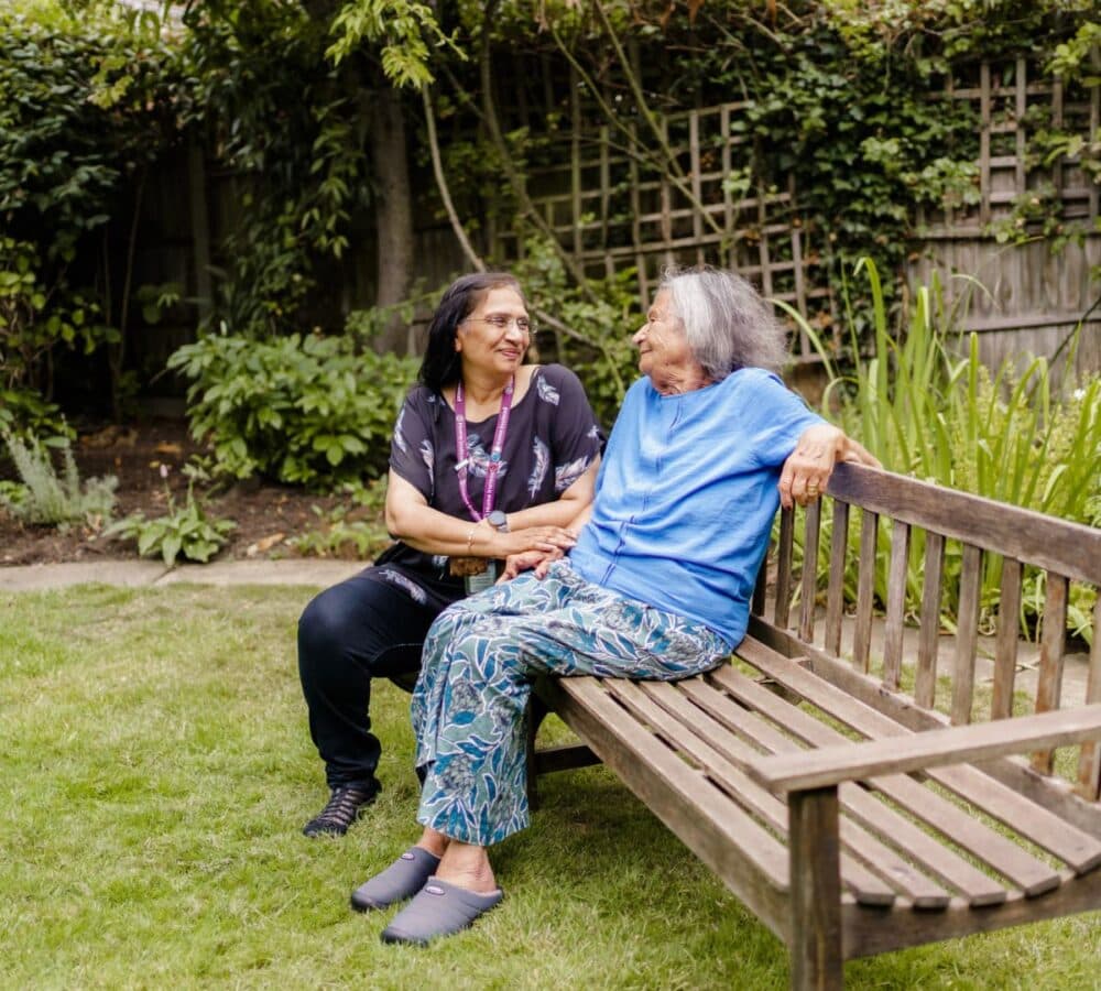A senior woman sitting on a bench with her Care Professional and having a chat with her while relaxing at her garden