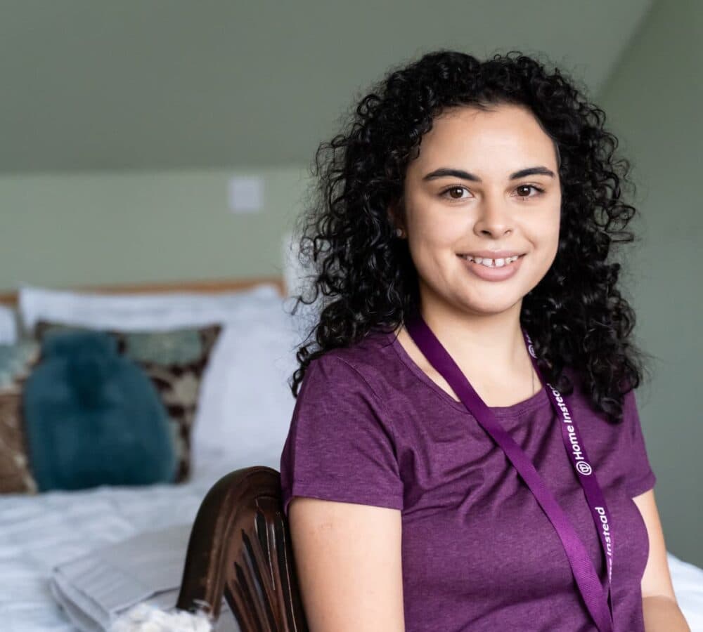 A young care professional with curly hair wearing an ID and wearing maroon type sitting inside a room