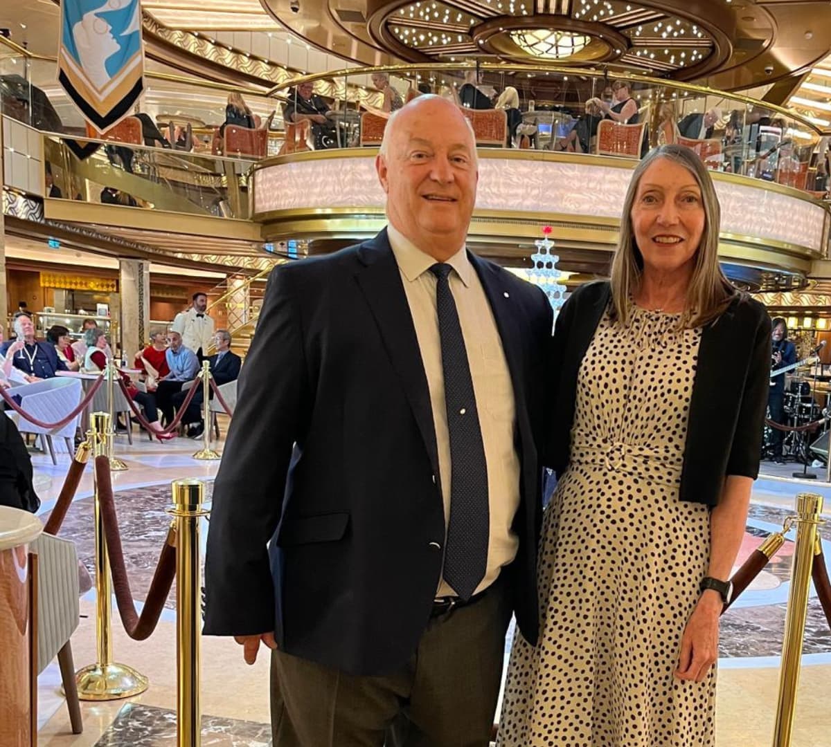 An old man wearing a suit and a necktie with a woman wearing a dress standing inside f a fancy restaurant with people and a piano in the background