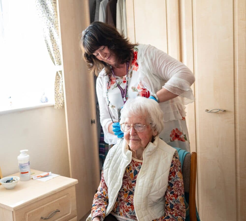 A carer with black hair and white top standing and wearing blue gloves while combing the hair of a senior wearing eyeglasses and with hair and sitting on a chair