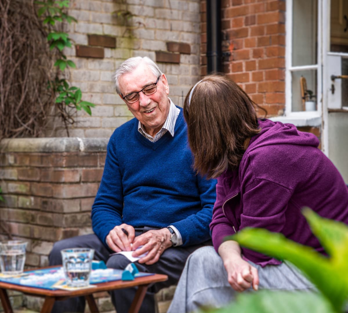 A Senior with a Care Professional wearing purple sweater outside the house drinking water and playing scrabble together