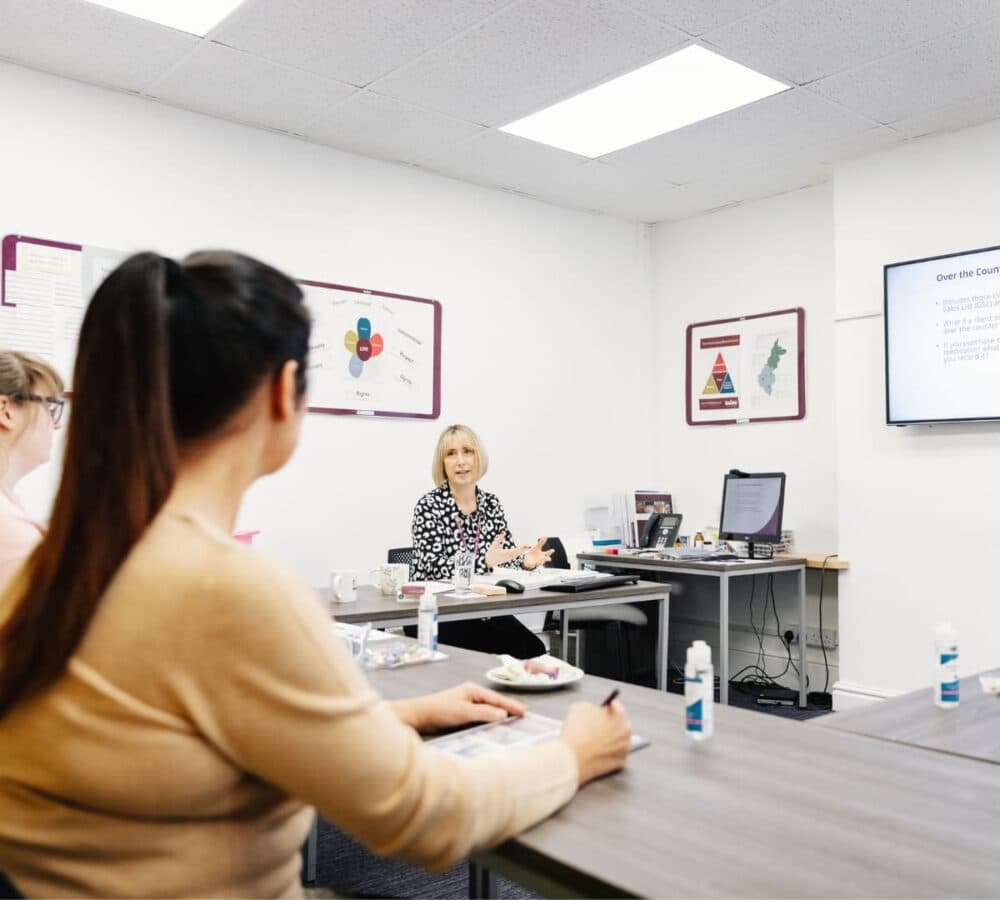 A woman teaching about over the counter medicine using a television with students sitting and with a table