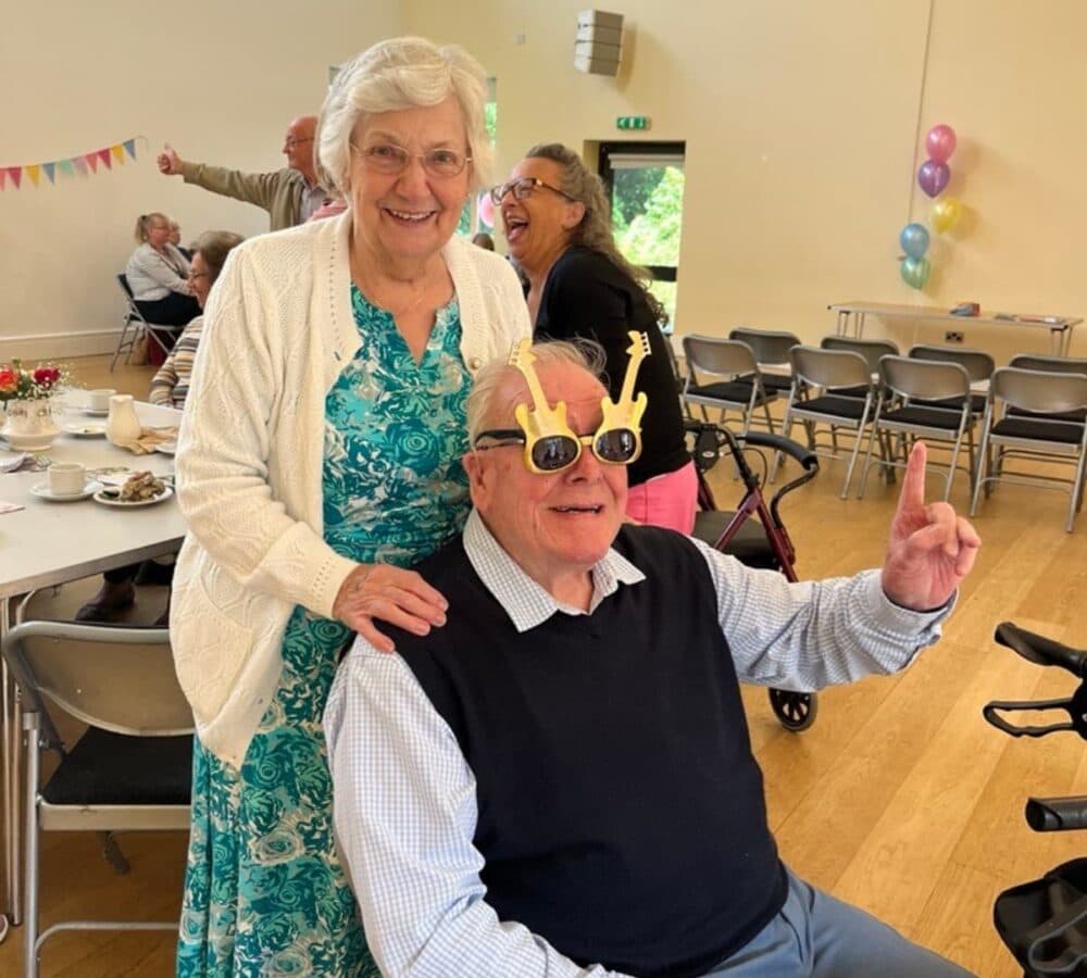 Elderly man wearing guitar-shaped glasses giving thumbs up, while a woman stands behind him, smiling. - Home Instead
