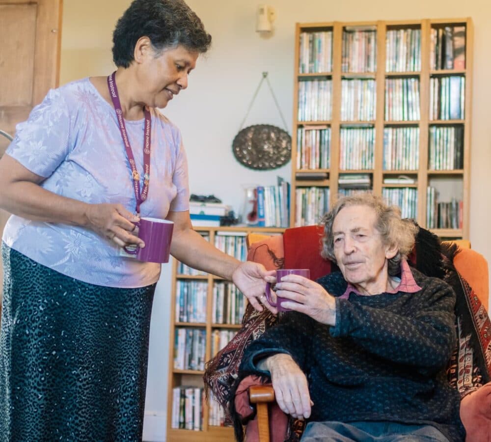 A senior male sitting on a couch inside a house and a carer wearing skirt giving a cup to the old man