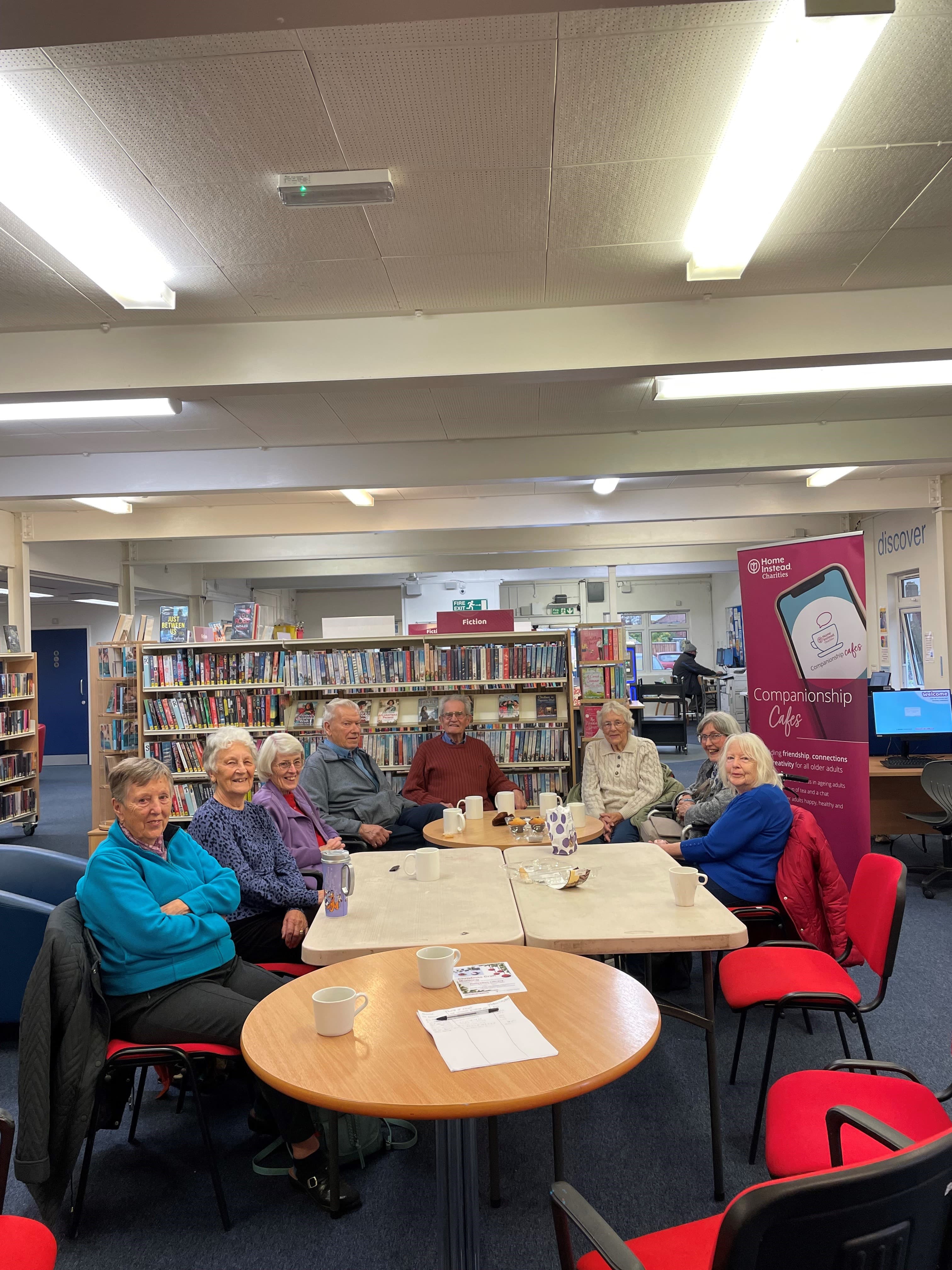 A group of older adults sit around tables with drinks in a library, chatting and smiling at the camera. - Home Instead